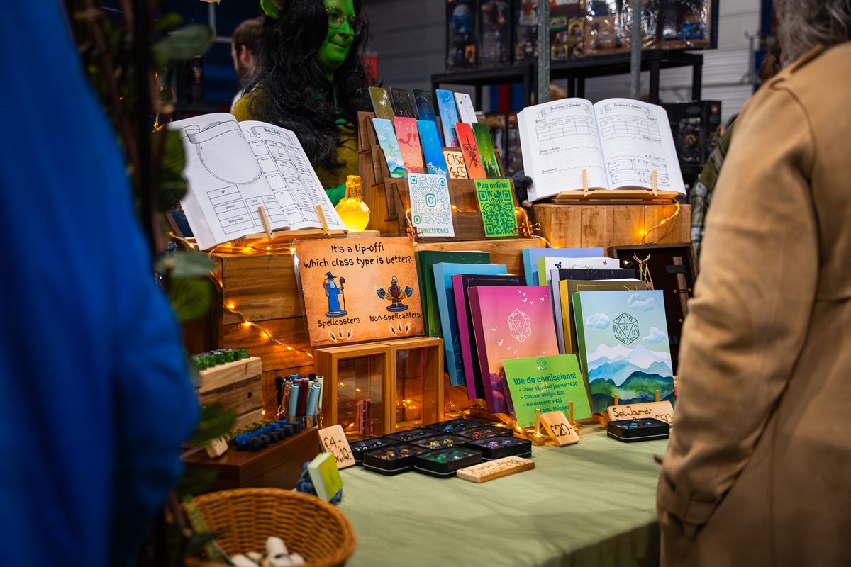 Artist-tafel op Con La Posta met handgemaakte journals, bookmarks en dice-sets, met een groen geschminkte verkoper