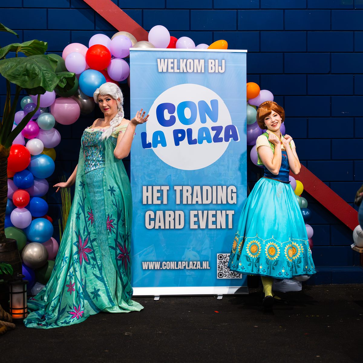 Elsa- en Anna-cosplayers poseren naast de welkomstbanner van Con La Posta, omringd door een ballonnenboog